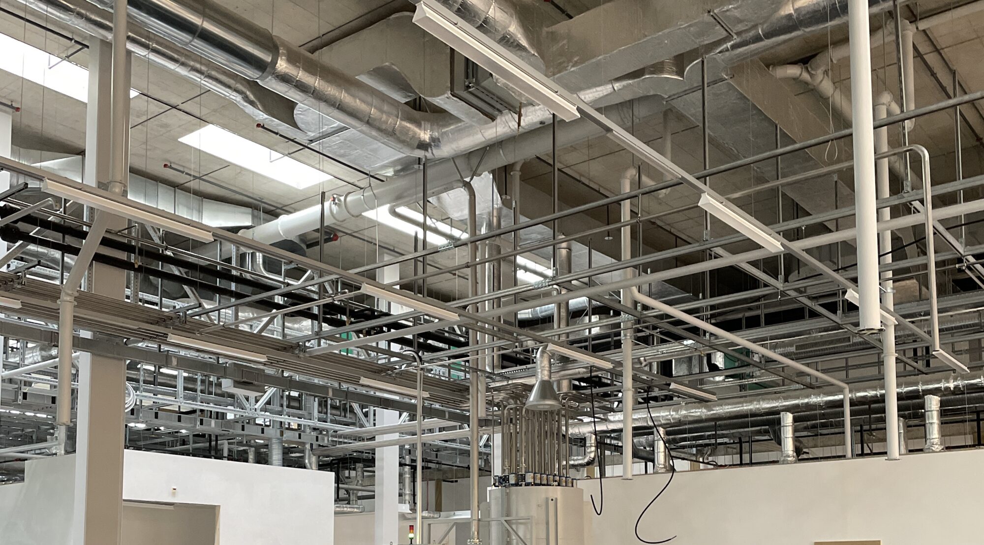 Halle de production à Neuchâtel : gaines de ventilation rectangulaires, réseaux de fluides et chemins de câbles en plafond technique au-dessus d’une ligne de production.